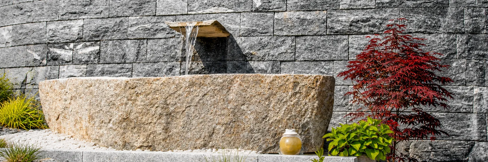 Wandbrunnen mit Wasserauslauf und großem Granittrog vor grauer Natursteinmauer, daneben roter japanischer Ahorn